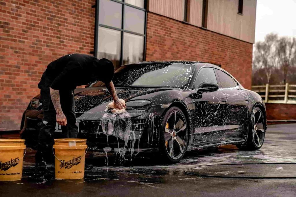 Professional detailer hand-washing a luxury black car with soap suds using the two-bucket method, highlighting the steps of exterior car detailing for a flawless, glossy finish.