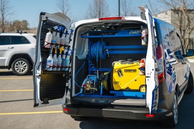 A fully-equipped mobile car detailing van parked in a lot in Frisco, Texas, showcasing organized shelves with cleaning chemicals, spray bottles, hoses, and a Champion generator, illustrating a modern on-site auto cleaning service ready for professional use.